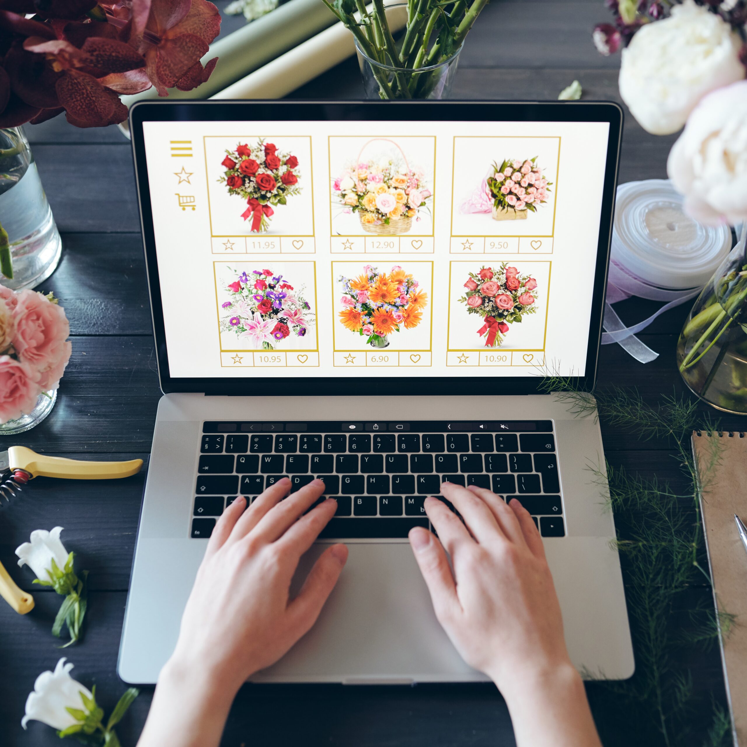 Above view of unrecognizable florist standing at desk with beautiful flowers and hand tools and typing on laptop keyboard