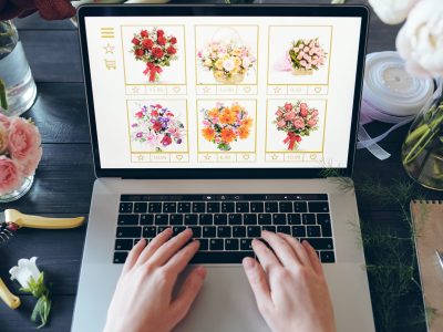 Above view of unrecognizable florist standing at desk with beautiful flowers and hand tools and typing on laptop keyboard